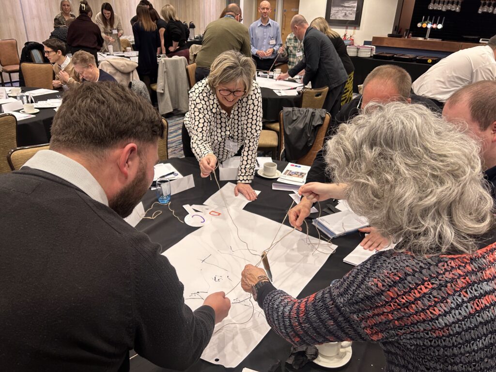 A group of people around a table, taking part in an ice breaker activity involving each person holding a piece of string that is attached to one pen between them. The pen is drawing on a piece of paper.